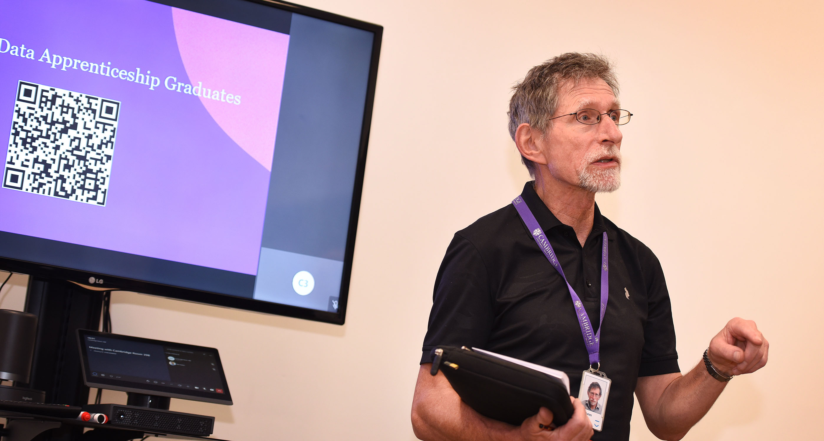 Tim Oates CBE faces outward and stands next to a screen displaying a QR code delivering a speech to the successful data apprentices and their line managers at the data apprentice celebration held at University Printing House in Cambridge.